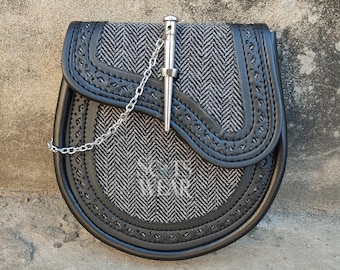 May include: A black leather sporran with a silver chain and a silver kilt pin. The sporran is decorated with a black and white herringbone pattern.