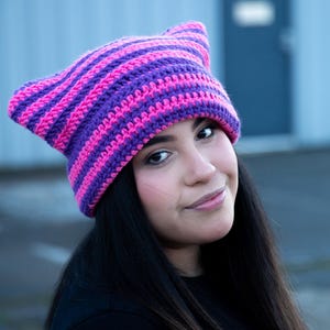 May include: A woman wearing a purple and pink striped crocheted hat with cat ears. The hat is made with a soft yarn and has a comfortable fit.