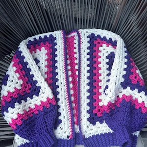 Peut inclure: Un cardigan en crochet fait à la main, sans fermeture, avec un motif de carrés granny. Le pull présente un design color block avec des carrés blancs, violets et roses. Les manches et les poignets sont violets unis, et le style général est décontracté et confortable.