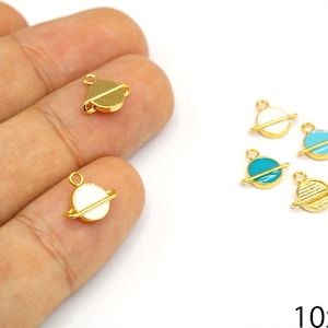 May include: Gold-colored planet charms with white and teal enamel details. The charms have a ring for attaching to jewelry. The image shows the charms in a hand and on a white surface. The charms are approximately 10x9mm.