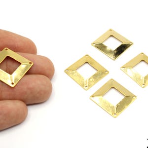 Könnte beinhalten: Vier goldfarbene rautenförmige Schmuckanhänger, jeweils mit einer quadratischen Öffnung in der Mitte und drei kleinen Löchern. Einer der Anhänger wird in einer Hand gehalten. Die Anhänger sind ca. 20 mm groß.