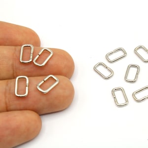 May include: A collection of small, silver-colored rectangular metal rings, approximately 10mm in size, displayed on a white background. Some rings are held in a hand, while others are scattered. These findings are suitable for jewelry making and crafts.