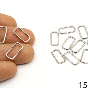 Puede incluir: Varios anillos rectangulares de metal plateado, cada uno con una pequeña abertura. Los anillos miden aproximadamente 1,5 cm de largo y 0,7 cm de ancho. Algunos se sostienen en una mano, otros están sobre una superficie blanca.