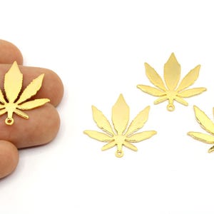 May include: Gold-colored metal leaf charms, resembling cannabis leaves, are displayed against a white background. One charm is held in a hand, while three others are arranged nearby. Each charm has a small loop for attaching to jewelry.