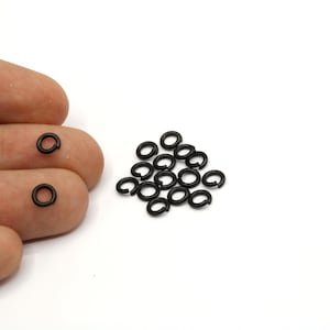 May include: A collection of small, matte black jump rings, some scattered and some held by fingers. The rings are circular and appear to be made of metal. The background is plain white.