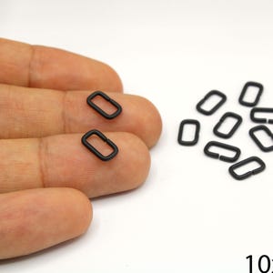 Puede incluir: Un conjunto de pequeños anillos metálicos negros de forma rectangular, algunos sobre una mano y otros dispersos sobre una superficie blanca. Los anillos miden aproximadamente 10 mm por 6 mm. Probablemente se utilizan para manualidades o joyería.