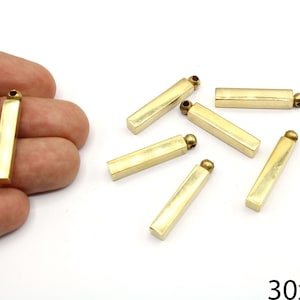 May include: Gold-colored rectangular metal charms with a small ball at one end. The charms are approximately 1.2 inches long and 0.2 inches wide. Several charms are scattered on a white surface.