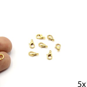 May include: Several small, gold-colored lobster claw clasps are scattered on a white surface. Two clasps are held by a hand. The clasps are approximately 5mm x 10mm in size. These clasps are used for jewelry making.