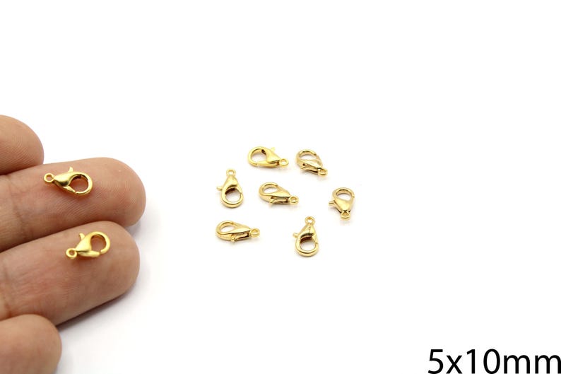 May include: Several small, gold-colored lobster claw clasps are scattered on a white surface. Two clasps are held by a hand. The clasps are approximately 5mm x 10mm in size. These clasps are used for jewelry making.