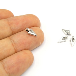Puede incluir: Pequeños dijes plateados con forma de flecha y acabado texturizado. Los dijes tienen forma triangular con un pequeño lazo en la parte superior para sujetarlos a la joyería. Se ven varios dijes.
