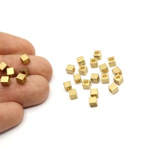 May include: A group of small, gold-colored metal cubes with a hole in the center. The cubes are scattered on a white surface. The cubes are approximately 4 mm in size.