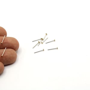 May include: A collection of small, silver-colored stud earrings with a flat, round, gold-colored top. Several earrings are scattered on a white surface, with two earrings held in a hand. The image includes the text "2mm".