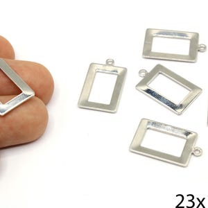 Peut inclure: Charm rectangulaire argenté avec une boucle pour la fabrication de bijoux. Les breloques mesurent environ 2,3 cm de long et 1,4 cm de large. Plusieurs breloques sont éparpillées sur une surface blanche, dont une tenue par une main.