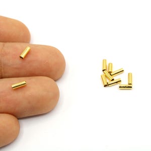May include: Small, gold-colored cylindrical beads. Several beads are scattered on a white surface, with a few resting on a person's fingers. The text "5mm" is visible in the lower right corner. These beads are likely for jewelry making.