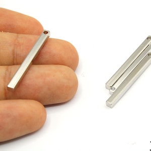 Puede incluir: Colgantes rectangulares plateados, cada uno con un pequeño agujero en un extremo. Un colgante se sostiene en una mano, mientras que otros tres están dispuestos sobre una superficie blanca. Incluye el texto "30mm".