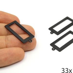 Puede incluir: Tres colgantes rectangulares negros de metal, con un lazo, ideales para la creación de joyas. Los colgantes miden 3,3 cm x 1,4 cm. Uno se sostiene en una mano, los otros dos sobre una superficie blanca.