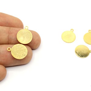 May include: Gold-colored, round metal charms with a textured surface and a small loop at the top for attaching to jewelry. The charms are approximately 0.5 inches in diameter. Several charms are shown, some held in a hand.