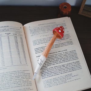 K&ouml;nnte beinhalten: Ein skurriler Stift mit roter Pilzkappe und wei&szlig;en Punkten liegt auf einem offenen Buch. Der Stiftschaft ist hellbraun, und das Wort "Judy" steht auf der wei&szlig;en Spitze. Das Buch zeigt wissenschaftlichen Text.