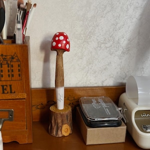 以下が含まれることがあります： 赤と白のドット柄の傘を持つ、キノコ型のペン。ペンの木製の茎は、小さく彫られた木の台座に置かれています。「Judy」という文字がペンに書かれています。ユニークなデスクアクセサリーです。