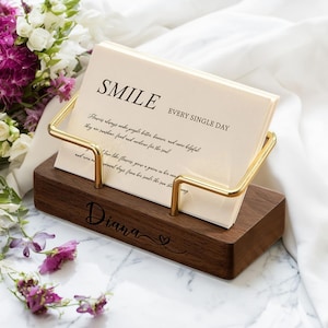 以下が含まれることがあります： 金色の金属フレームが付いた木製の卓上カレンダーホルダー。「SMILE」という単語と「EVERY SINGLE DAY」というフレーズが表示されています。ベースには「Diana」という名前とハートのシンボルが刻まれています。背景には花があります。