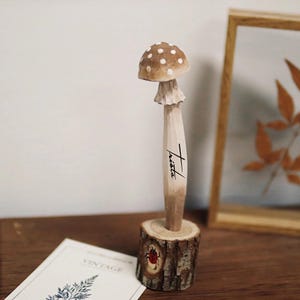 K&ouml;nnte beinhalten: Eine Holzpilzskulptur mit braunem Hut und wei&szlig;en Punkten, montiert auf einem kleinen Holzscheibenfu&szlig;. Der Stiel ist hellbraun mit dem Wort "Trista" in schwarzer Schrift. Ein kleiner roter Marienk&auml;fer ist in die Basis geschnitzt.