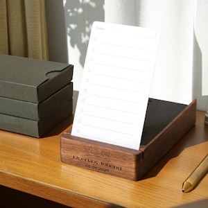 Peut inclure: Un organiseur de bureau en bois avec un bloc-notes blanc. Le bloc-notes comporte un en-tête "Today" et des lignes numérotées. L'organisateur est gravé avec "Liceria & Johnny" et la date "20.05.2025". Une pile de boîtes vert foncé est visible en arrière-plan.