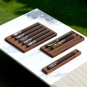 May include: Several black pens with silver accents and the text "TEST 2" are displayed in wooden trays. One tray holds a pen with gold detailing. The trays are on a white marble surface.