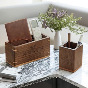Peut inclure: Deux organiseurs de bureau en bois avec texte gravé et motif floral. L'un contient une télécommande et un carnet en cuir. L'autre contient des stylos. Le texte se lit comme suit : "Mr & Mrs Small, Merci d'avoir fait partie de notre célébration ! 30 janvier 2024."