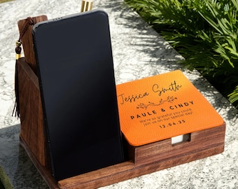 May include: A wooden phone stand with a built-in notepad. The stand holds a black smartphone and an orange notepad with the names "Jessica Smith" and "Paule & Cindy" and the date "12.04.25" printed on it.