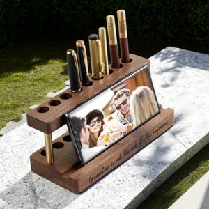 Peut inclure: Un organiseur de bureau en bois avec un support pour téléphone et des emplacements pour stylos. L'organisateur contient plusieurs stylos et un smartphone affichant une photo. La phrase "Graduation is not the end, it's the beginning" est gravée sur la base.