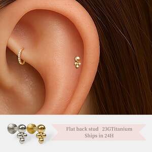 May include: A close-up of a gold flat back stud earring with three small balls. The earring is shown in a person's ear. The text "Flat back stud 23G Titanium Ships in 24H" is visible below the image.