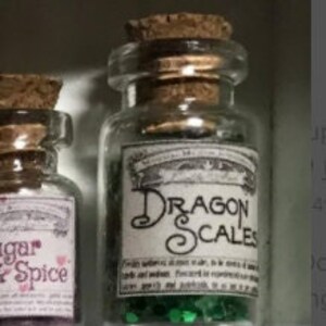 May include: A small, clear glass bottle with a cork stopper and a label that reads "Dragon Scales." The bottle contains green, glittery material. The label has decorative scrollwork and additional text.