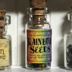 Könnte beinhalten: Kleine Glasflaschen mit Korken. Die mittlere Flasche hat ein Etikett mit der Aufschrift "RAINBOW SEEDS" über einem Regenbogenverlauf. Die Flasche enthält bunte Samen. Andere Flaschen haben ähnliche Designs.