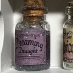 Könnte beinhalten: Kleine, klare Glasflasche mit Korken und lila Etikett mit der Aufschrift "Dreaming Draught". Das Etikett hat dekorative Sterne und Wirbel. Die Flasche ist mit einer schimmernden, dunklen Flüssigkeit gefüllt.
