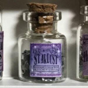 May include: A small, clear glass bottle with a cork stopper and a purple label reading "STARDUST". The bottle contains dark granules. The label has a vintage design. The bottle is part of a collection.