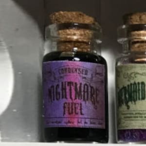 Könnte beinhalten: Kleine Glasflasche mit Korken und lila Etikett mit der Aufschrift "NIGHTMARE FUEL". Auf dem Etikett steht auch "CONDENSED". Die Flasche enthält eine dunkle Flüssigkeit. Die Flasche steht auf einem Regal.