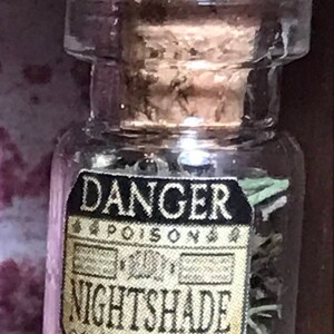 May include: A small, clear glass bottle with a cork stopper. The label reads "DANGER POISON NIGHTSHADE" in black and gold lettering. The bottle contains dried herbs.