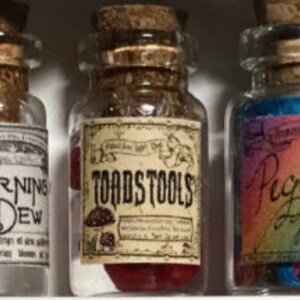 Op de afbeelding: Drie kleine glazen flessen met kurk stoppers en etiketten. De middelste fles leest "TOADSTOOLS" met een paddenstoel illustratie. De andere flessen hebben etiketten met de tekst "MORNING DEW" en "Pegasus Tears". De flessen zijn gevuld met kleurrijke inhoud.