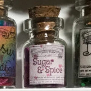 Op de afbeelding: Drie kleine glazen flesjes met kurken stoppen. Het middelste flesje heeft een etiket met de tekst "Sugar & Spice". De flesjes bevatten kleurrijke inhoud. De flesjes worden op een witte plank tentoongesteld.