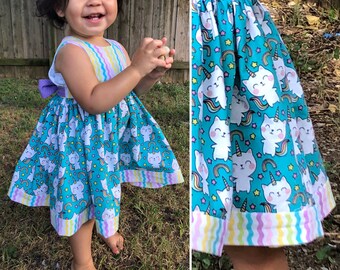 Può includere: Abito turchese senza maniche con stampa gatto-unicorno, arcobaleni e stelle. L'abito ha un corpetto e un orlo a righe colorate e un fiocco viola sul retro. L'abito è progettato per un bambino.