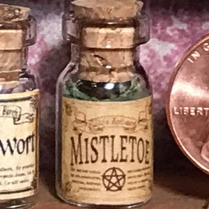 Könnte beinhalten: Eine kleine Glasflasche mit Korken, beschriftet mit "Mistletoe". Das Etikett ist gealtert und zeigt ein Pentagramm-Symbol. Die Flasche enthält eine grüne Flüssigkeit. Daneben eine weitere Flasche mit der Aufschrift "Bugwort".