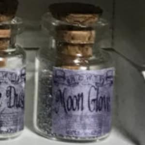 Könnte beinhalten: Kleine Glas-Zaubertrankflasche mit Korken und lila Etikett mit der Aufschrift "Moon Glow". Die Flasche ist mit einer schimmernden, silbrigen Substanz gefüllt. Die Flasche steht auf einem weißen Regal.