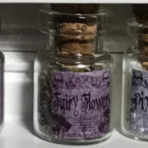 Könnte beinhalten: Drei kleine Glasflaschen mit Korken. Jede Flasche hat ein lila Etikett mit Text. Die mittlere Flasche trägt die Aufschrift "Fairy Flowers". Die Flaschen sind mit einer Substanz gefüllt, möglicherweise Glitzer oder getrockneten Blumen.