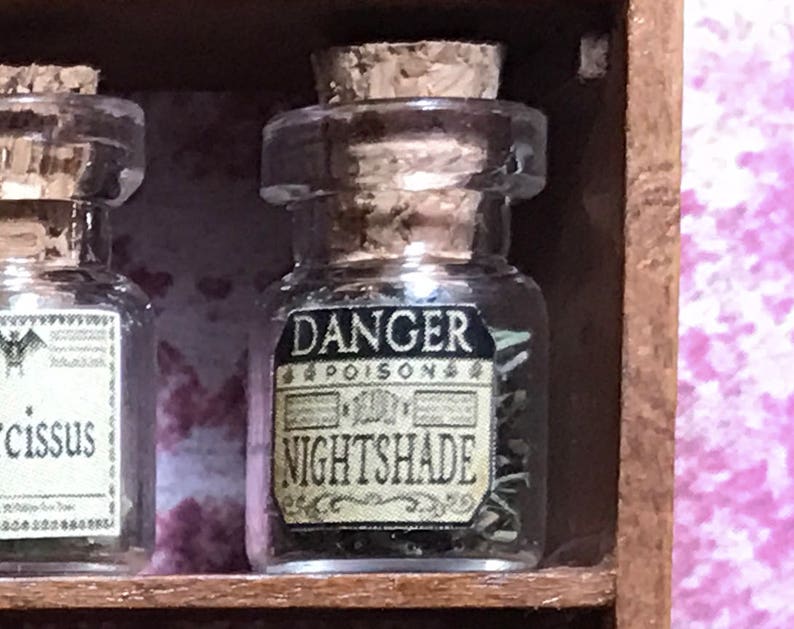 May include: A small, clear glass apothecary jar with a cork stopper. The label reads "DANGER POISON NIGHTSHADE" in black and cream. The jar is filled with green dried herbs. The jar is displayed on a wooden shelf.