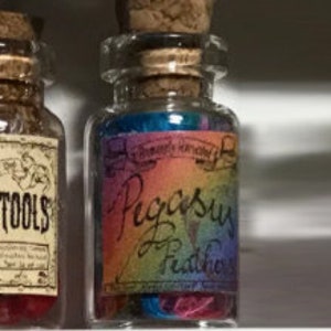 Op de afbeelding: Een kleine glazen fles met een kurkstop en een kleurrijk etiket met de tekst "Pegasus Feathers". Het etiket heeft een regenboogverloop en de fles bevat een mix van kleurrijke inhoud. De fles staat op een plank.