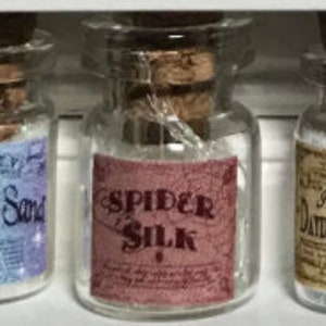 Op de afbeelding: Drie kleine glazen flesjes met kurken stoppen. De middelste fles heeft een etiket met de tekst "SPIDER SILK" in rode tekst. De andere flessen hebben etiketten met de teksten "Spring Sand" en "Daydream".