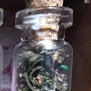 May include: A small, clear glass jar with a cork stopper, filled with dried green herbs and a small green object. The jar is set against a dark wooden background, suggesting a natural or apothecary theme.