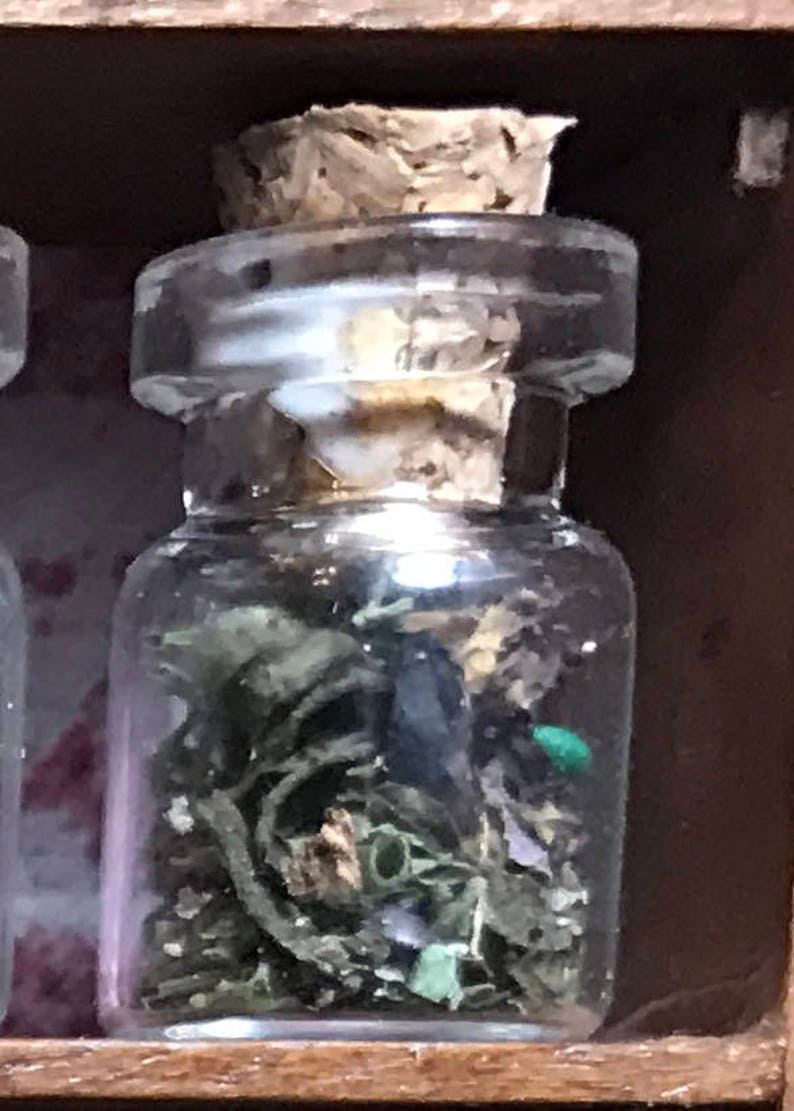 May include: A small, clear glass jar with a cork stopper, filled with dried green herbs and a small green object. The jar is set against a dark wooden background, suggesting a natural or apothecary theme.