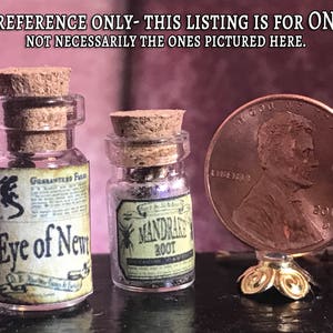 May include: Two small glass jars with cork stoppers, labeled "Eye of Newt" and "Mandrake Root." A penny is included for size reference. The jars have vintage-style labels with illustrations. The text "SIZE REFERENCE ONLY" is at the top.