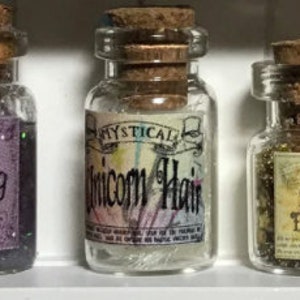 Könnte beinhalten: Drei kleine Glasflaschen mit Korken werden gezeigt. Die mittlere Flasche hat ein Etikett mit der Aufschrift "Mystical Unicorn Hair". Die Flaschen enthalten Glitzer und andere Materialien. Die Flaschen stehen auf einem weißen Regal.